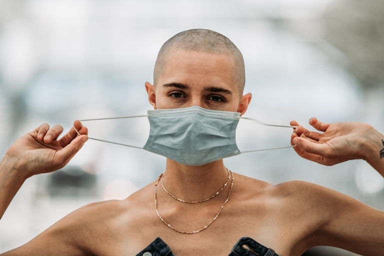 A bald woman wearing a thin necklace is putting on a blue surgical mask, looking directly at the camera.